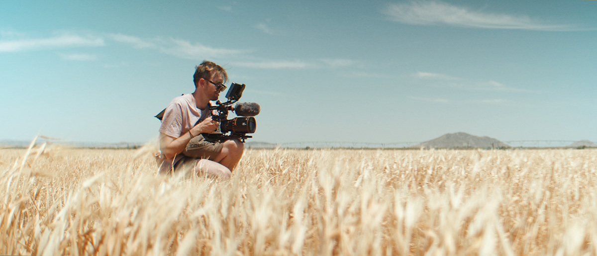 Max kneels in a field holding a camera