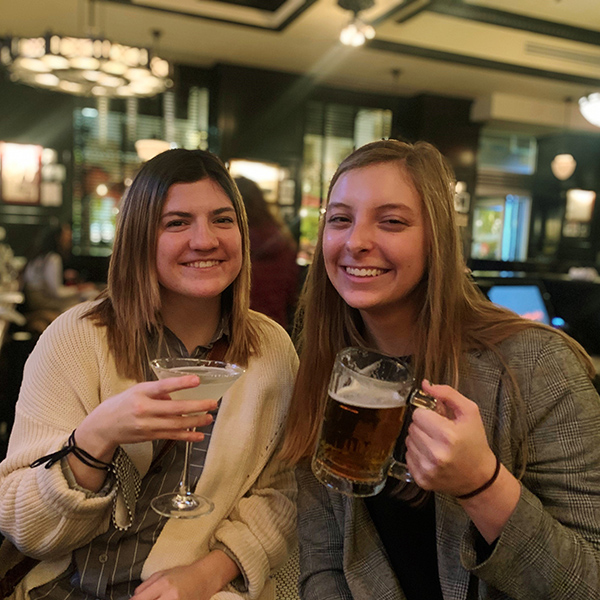 Lauren Hofstra and Cami Mondeaux clinking beer mugs together in pub